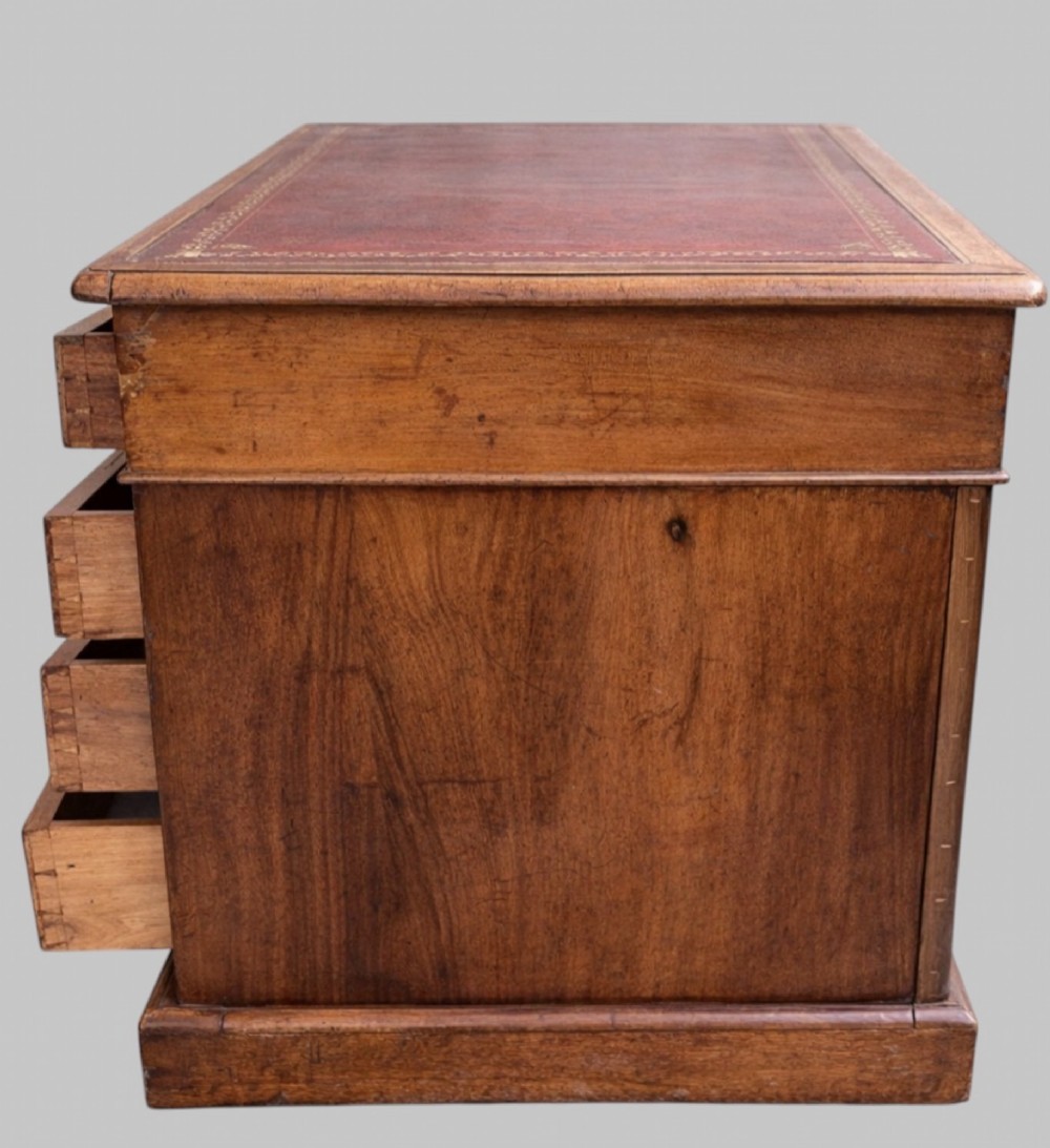 A late Victorian mahogany pedestal desk featuring an inset red leather writing surface, nine drawers with round brass pulls, and two plinth bases. View VI