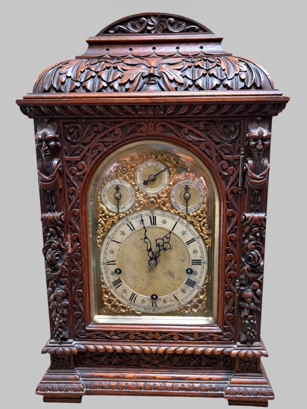 An intricately carved dark oak bracket clock featuring an arched brass dial, corner mask carvings, and foliate cresting.