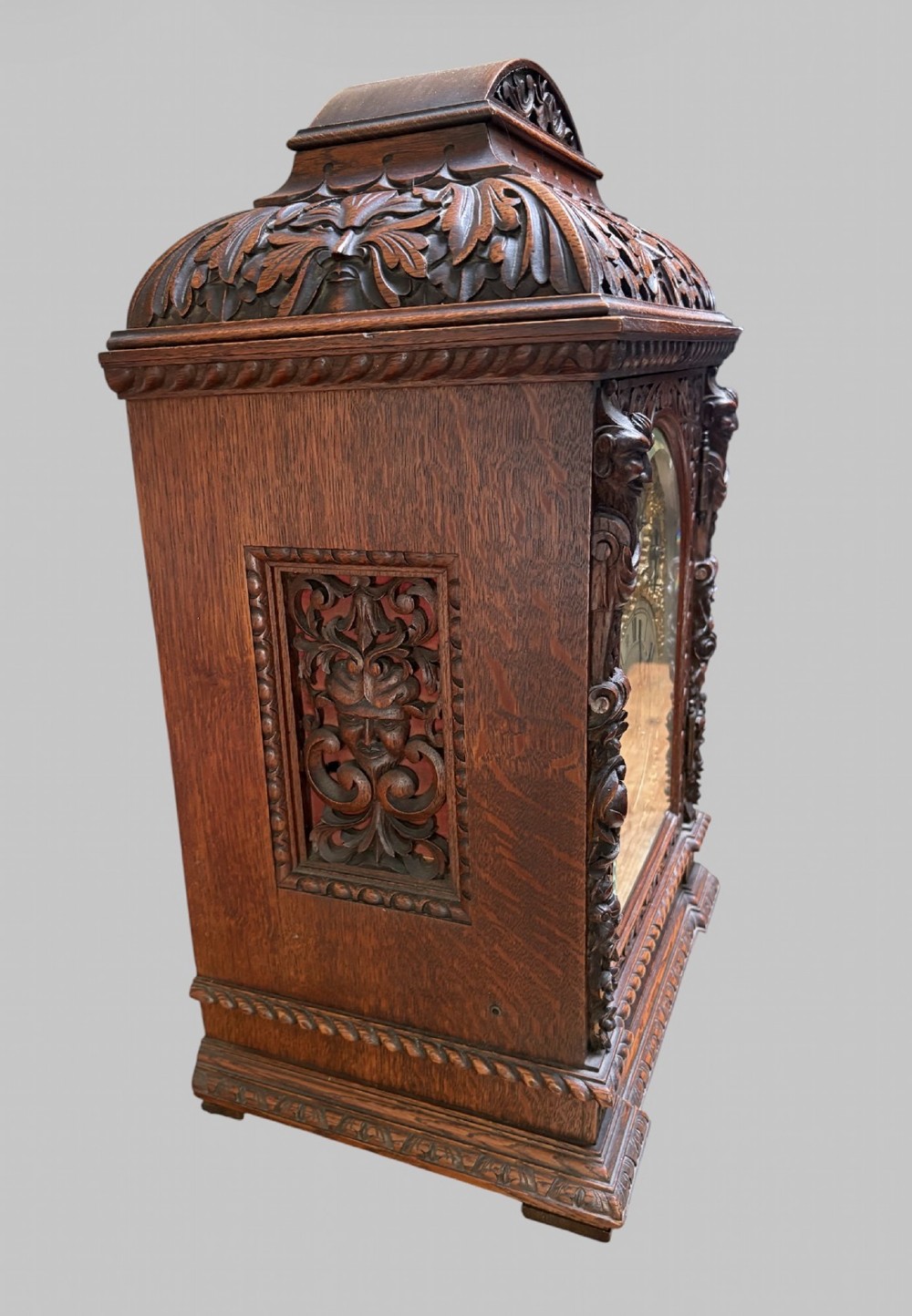 An intricately carved dark oak bracket clock featuring an arched brass dial, corner mask carvings, and foliate cresting. View II