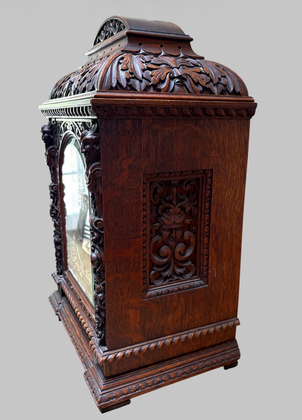 An intricately carved dark oak bracket clock featuring an arched brass dial, corner mask carvings, and foliate cresting. View III