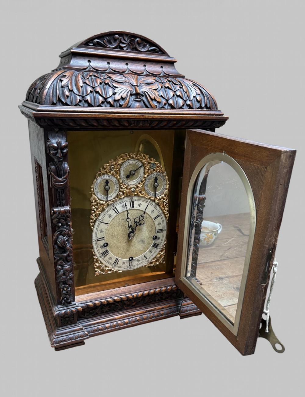 An intricately carved dark oak bracket clock featuring an arched brass dial, corner mask carvings, and foliate cresting. View V