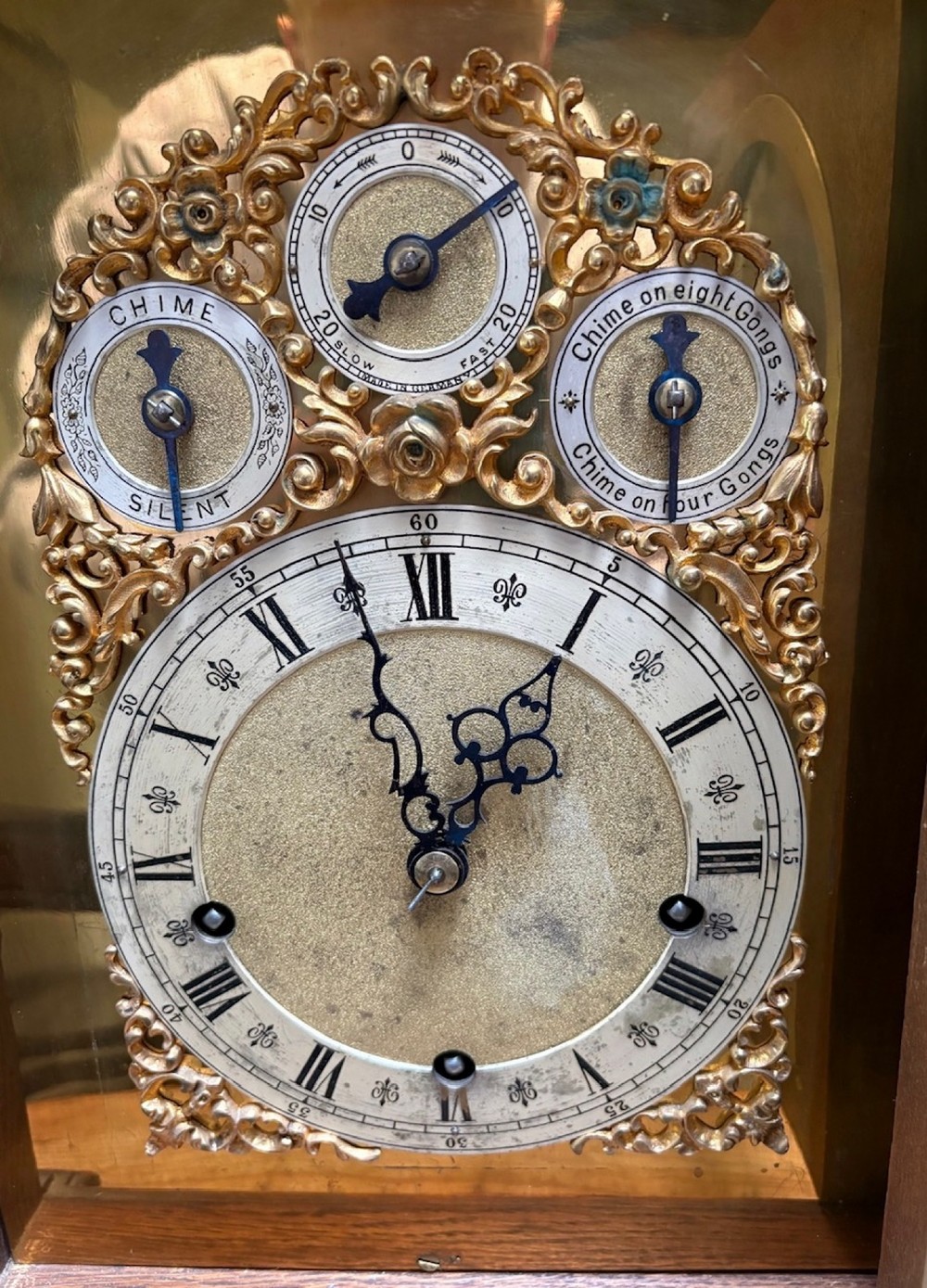 An intricately carved dark oak bracket clock featuring an arched brass dial, corner mask carvings, and foliate cresting. View VI