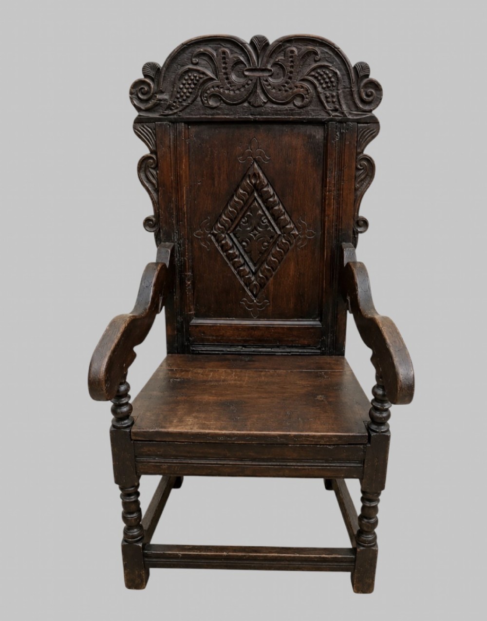 A dark carved oak wainscot armchair featuring a detailed cresting rail, central lozenge panel back, curved arms, and turned front legs.