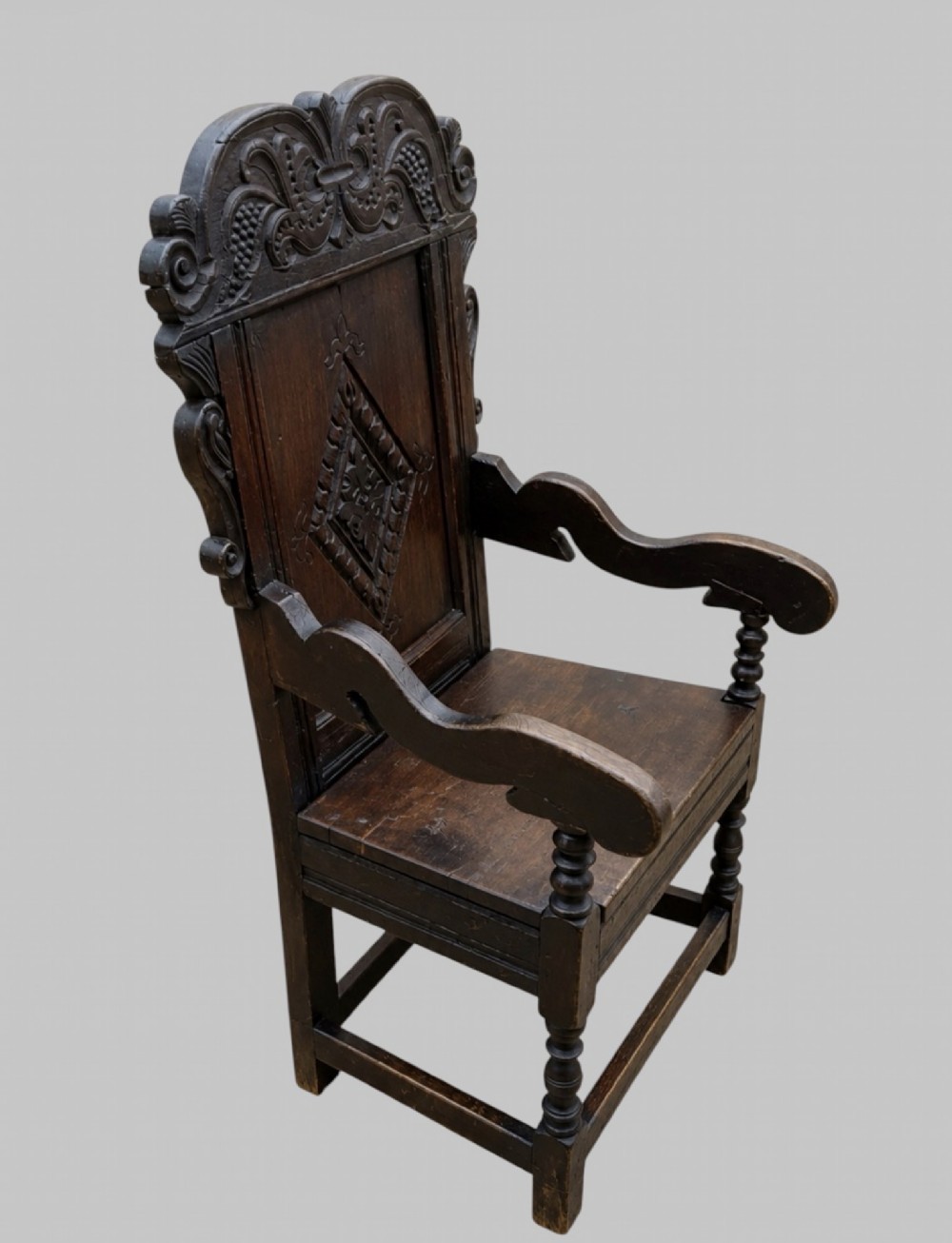 A dark carved oak wainscot armchair featuring a detailed cresting rail, central lozenge panel back, curved arms, and turned front legs. View II
