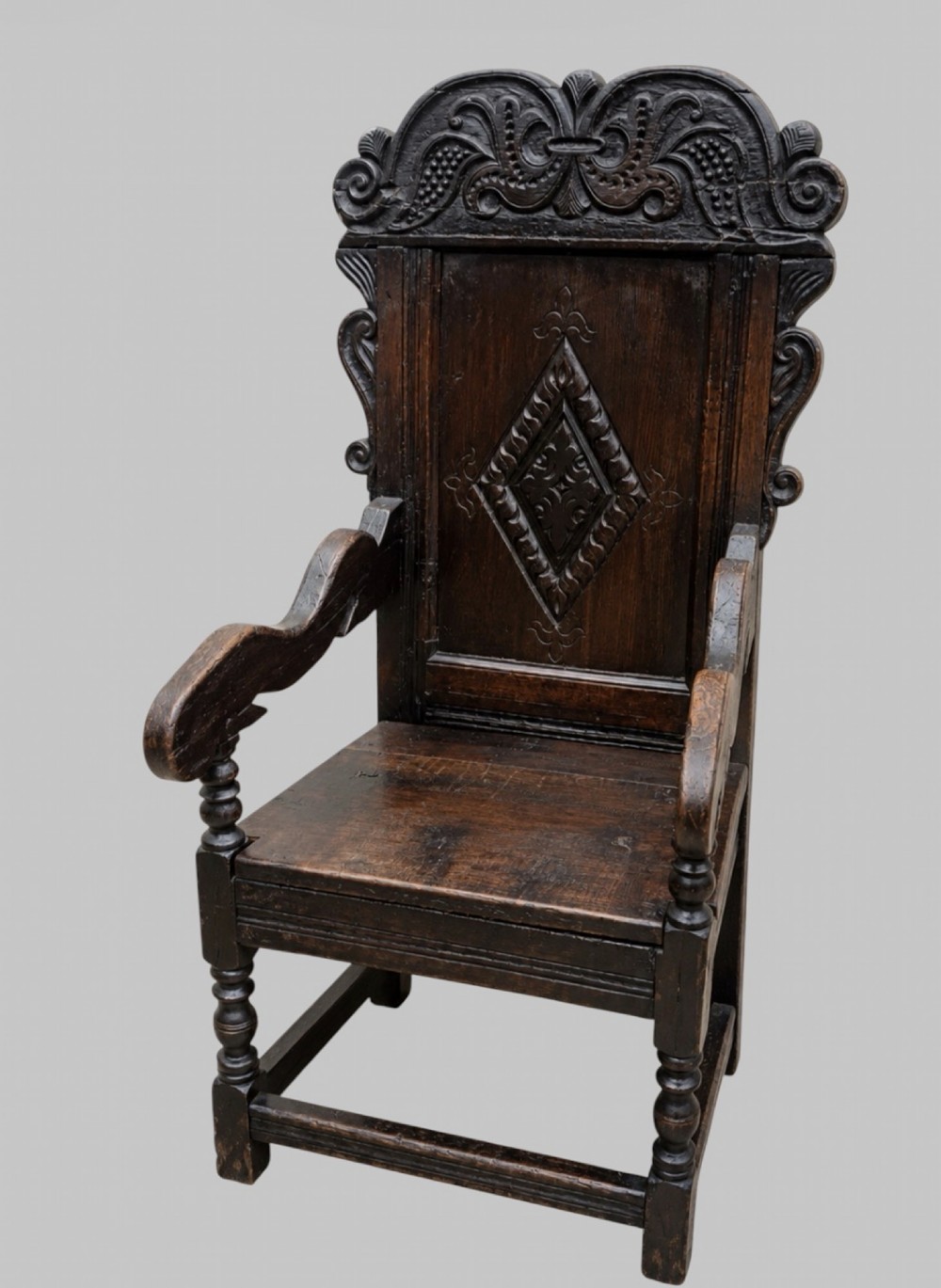 A dark carved oak wainscot armchair featuring a detailed cresting rail, central lozenge panel back, curved arms, and turned front legs. View III