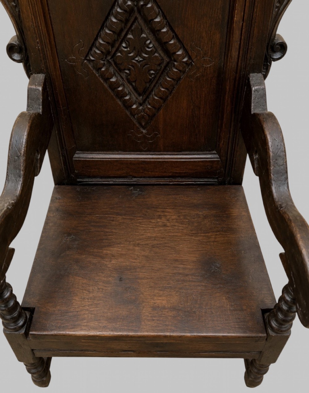 A dark carved oak wainscot armchair featuring a detailed cresting rail, central lozenge panel back, curved arms, and turned front legs. View V