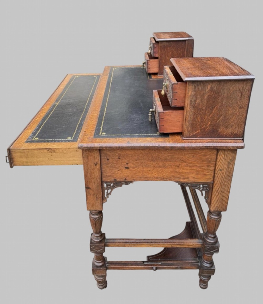 Victorian carved oak writing desk featuring a leather inset surface, two raised drawer cabinets on the top, two carved frieze drawers with brass drop handles, turned legs, and stretchers. View VI
