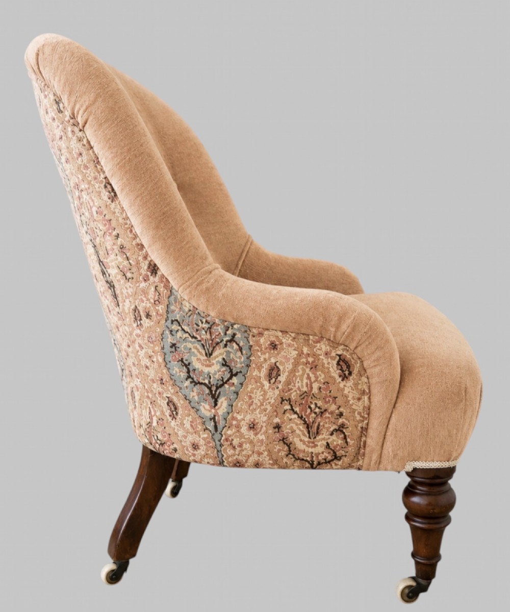 A profile view of a low Victorian iron-back nursing chair upholstered in beige velvet with printed pattern fabric sides and turned wooden legs on castors.
