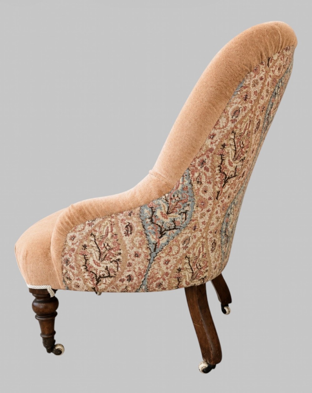 A profile view of a low Victorian iron-back nursing chair upholstered in beige velvet with printed pattern fabric sides and turned wooden legs on castors. View III