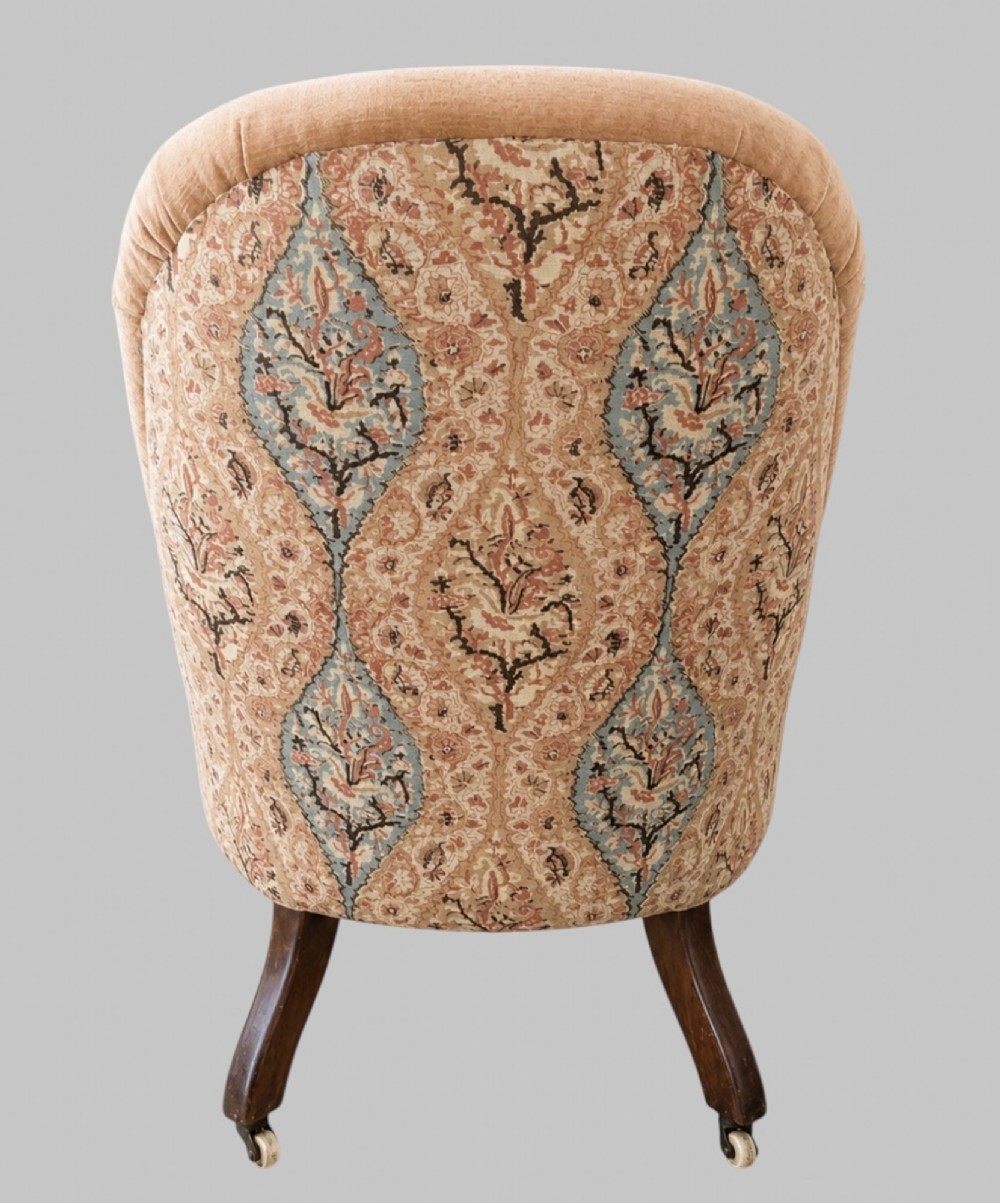 A profile view of a low Victorian iron-back nursing chair upholstered in beige velvet with printed pattern fabric sides and turned wooden legs on castors. View IV