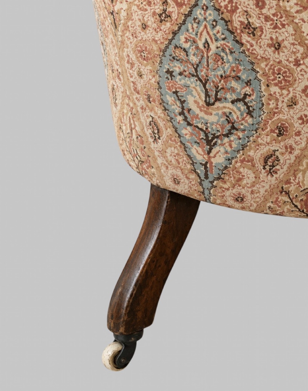 A profile view of a low Victorian iron-back nursing chair upholstered in beige velvet with printed pattern fabric sides and turned wooden legs on castors. View VI