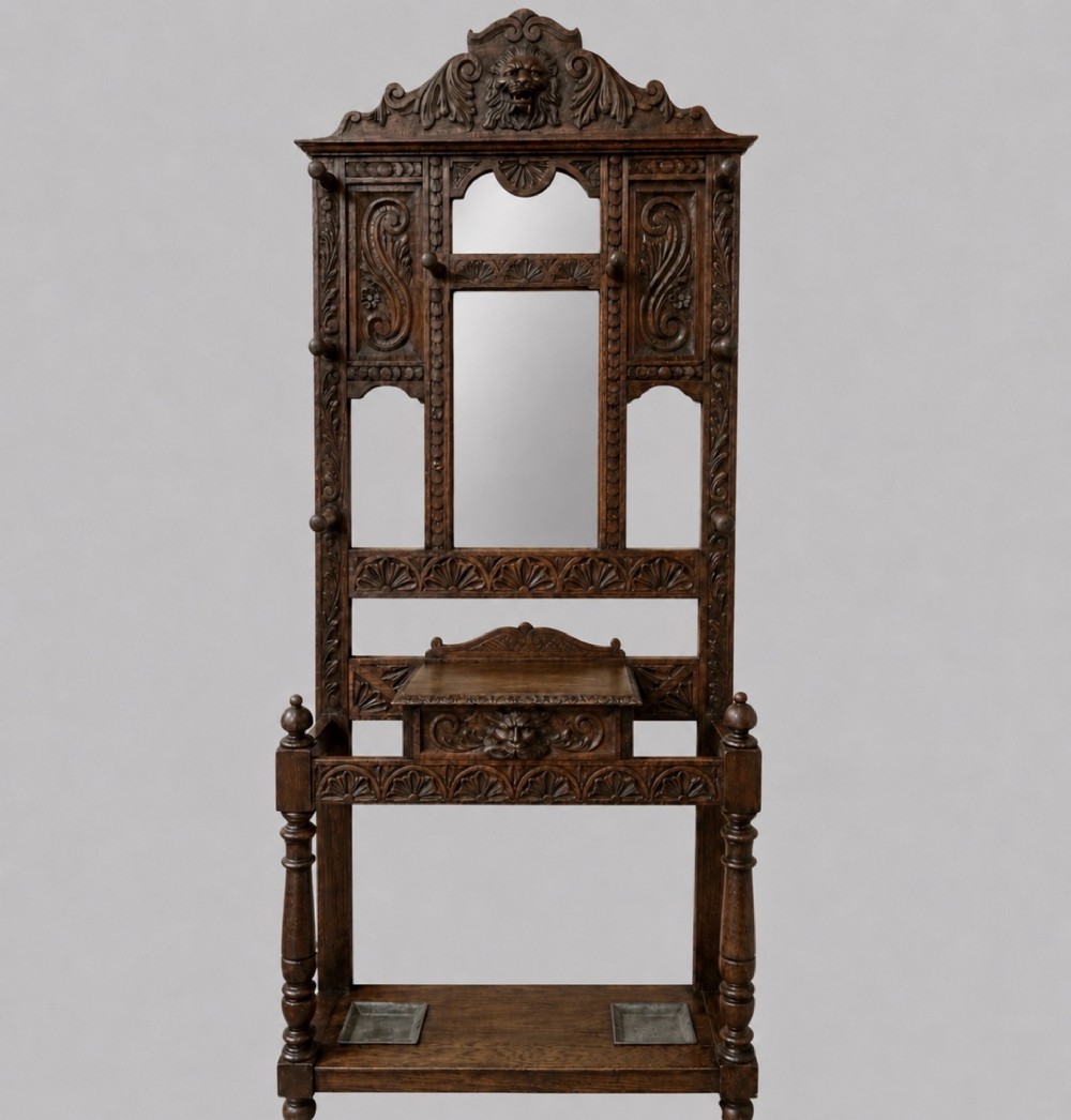 An antique carved dark oak hall stand featuring a central mirror, relief Green Man carved face motifs, hanging pegs, a middle drawer, and an umbrella rack base with metal drip trays.