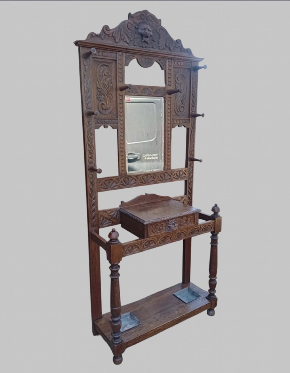 An antique carved dark oak hall stand featuring a central mirror, relief Green Man carved face motifs, hanging pegs, a middle drawer, and an umbrella rack base with metal drip trays. View II