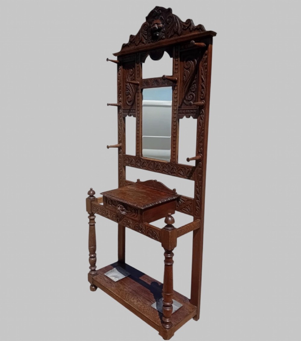 An antique carved dark oak hall stand featuring a central mirror, relief Green Man carved face motifs, hanging pegs, a middle drawer, and an umbrella rack base with metal drip trays. View III