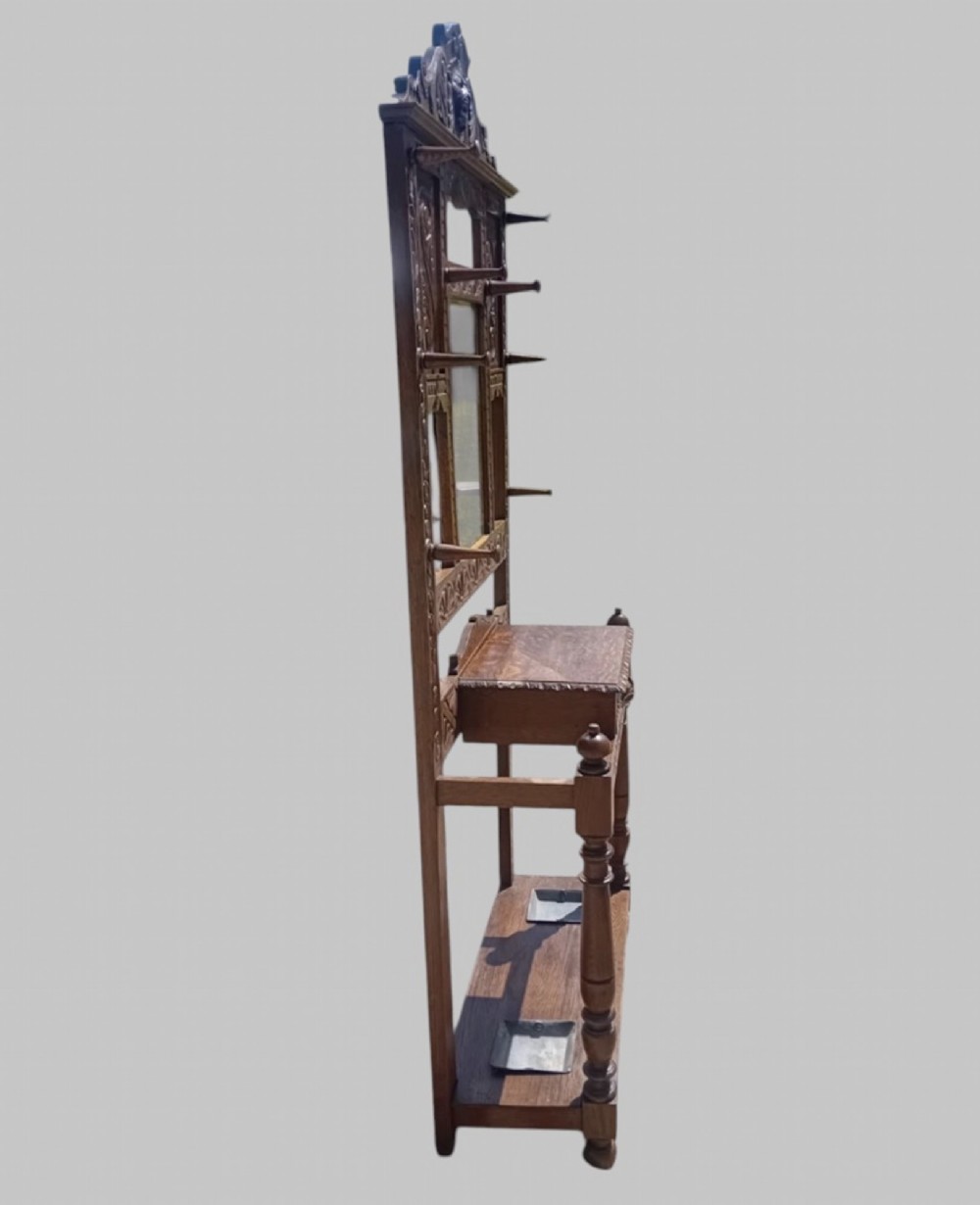 An antique carved dark oak hall stand featuring a central mirror, relief Green Man carved face motifs, hanging pegs, a middle drawer, and an umbrella rack base with metal drip trays. View IV