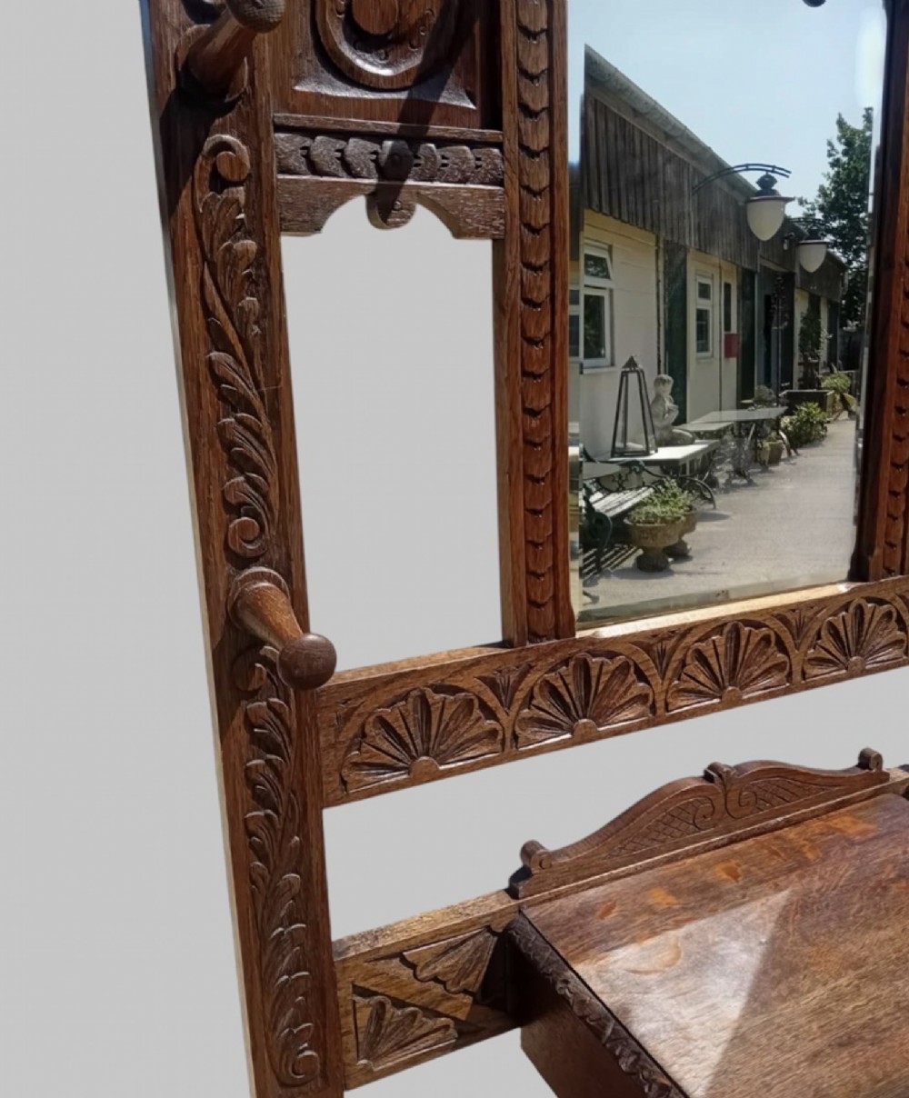 An antique carved dark oak hall stand featuring a central mirror, relief Green Man carved face motifs, hanging pegs, a middle drawer, and an umbrella rack base with metal drip trays. View V