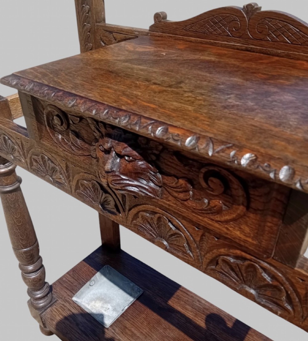 An antique carved dark oak hall stand featuring a central mirror, relief Green Man carved face motifs, hanging pegs, a middle drawer, and an umbrella rack base with metal drip trays. View VII