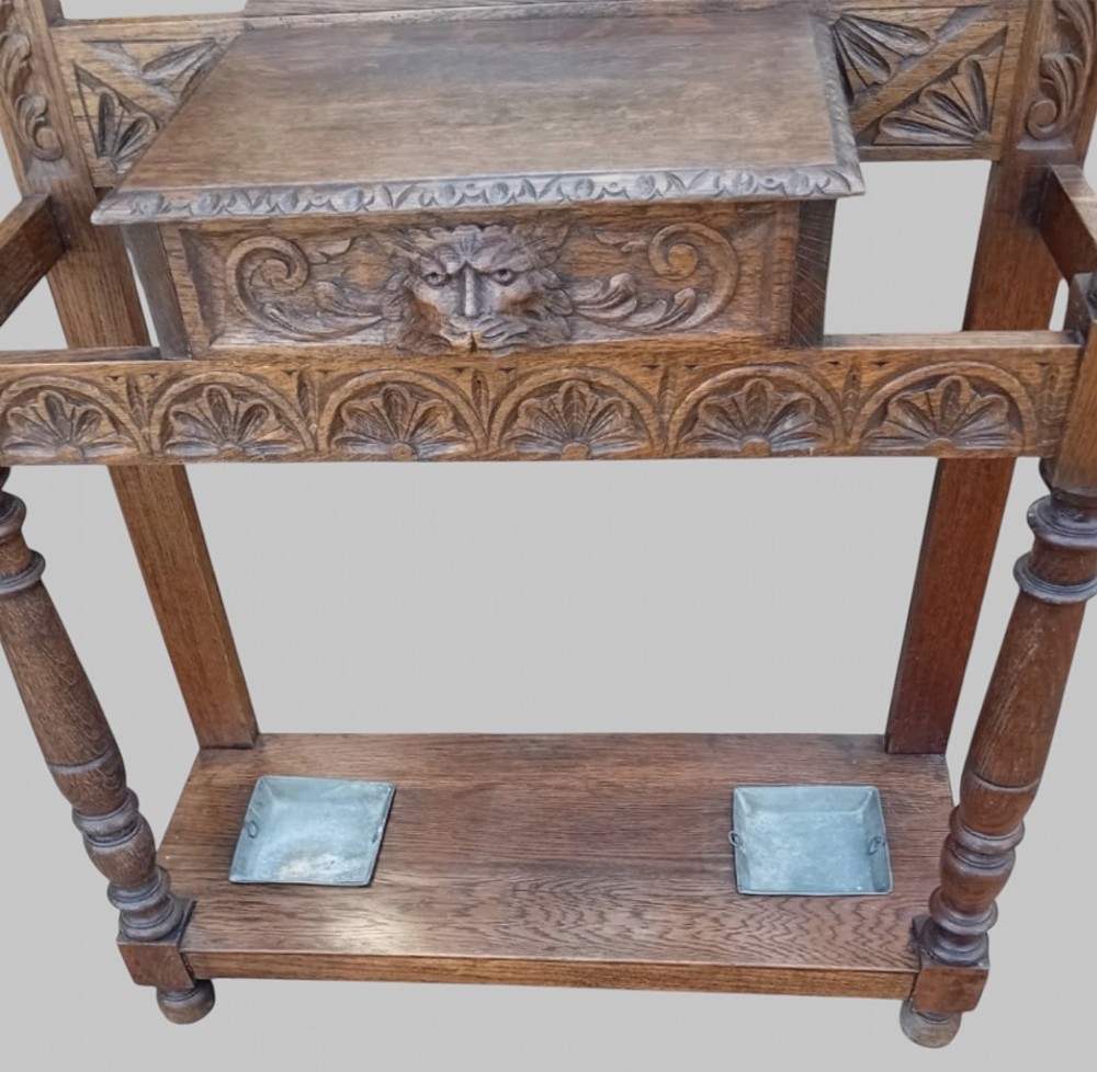 An antique carved dark oak hall stand featuring a central mirror, relief Green Man carved face motifs, hanging pegs, a middle drawer, and an umbrella rack base with metal drip trays. View VIII