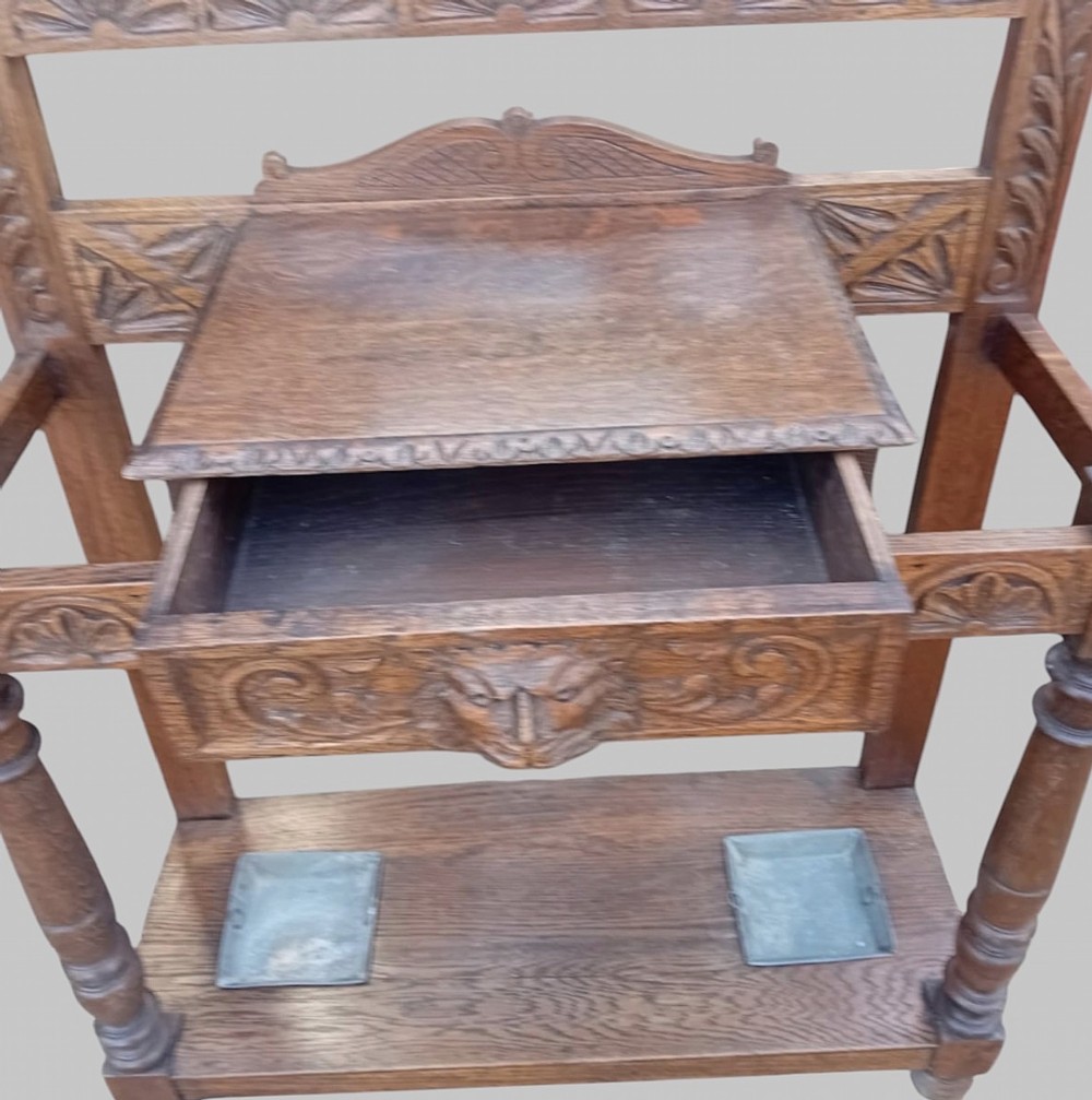 An antique carved dark oak hall stand featuring a central mirror, relief Green Man carved face motifs, hanging pegs, a middle drawer, and an umbrella rack base with metal drip trays. View IX