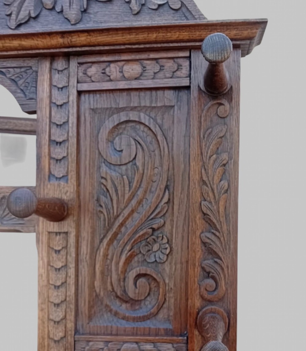 An antique carved dark oak hall stand featuring a central mirror, relief Green Man carved face motifs, hanging pegs, a middle drawer, and an umbrella rack base with metal drip trays. View X
