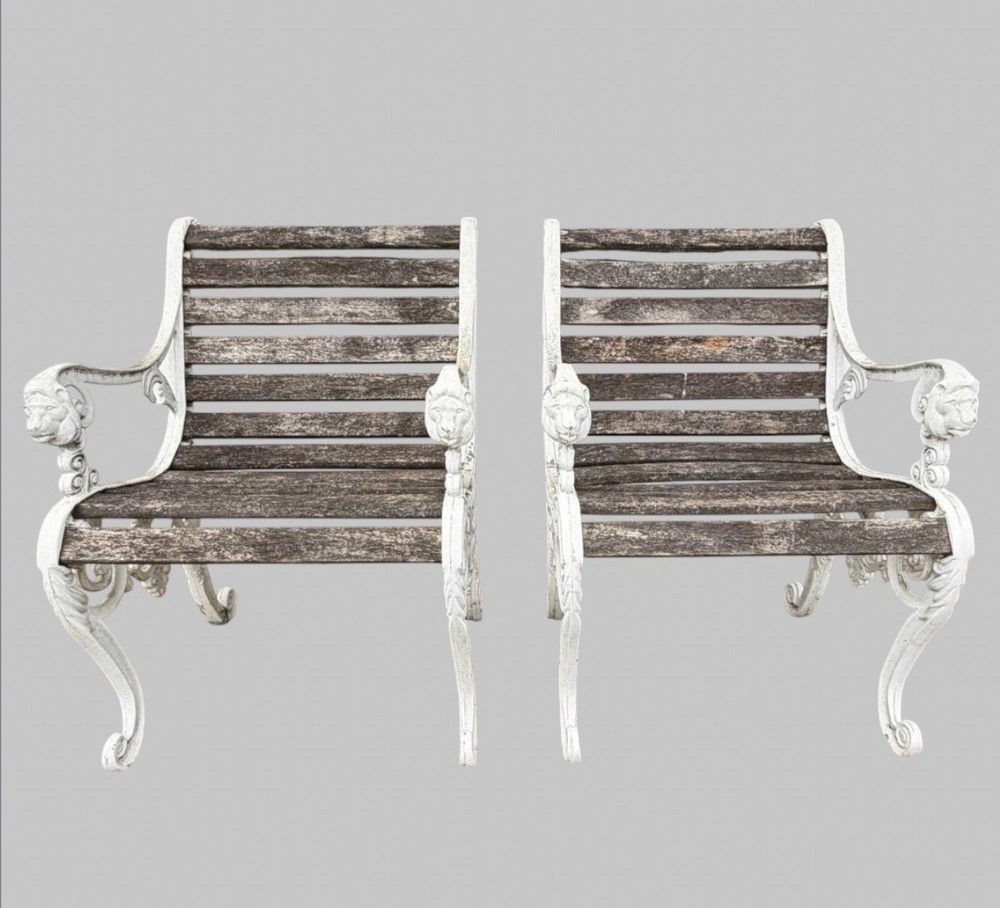 A pair of cast iron garden armchairs featuring white painted frames with lion mask armrest terminals and weathered timber slats.