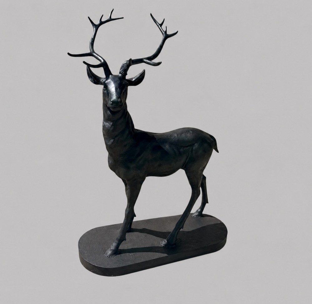 A patinated dark bronze sculpture depicting a standing young stag with antlers fixed onto an oval base.