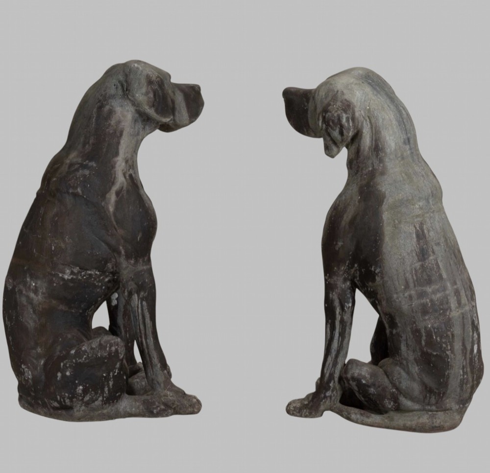 A pair of life-size cast lead sculptures depicting seated hounds with detailed anatomical features and a weathered grey surface. View IV