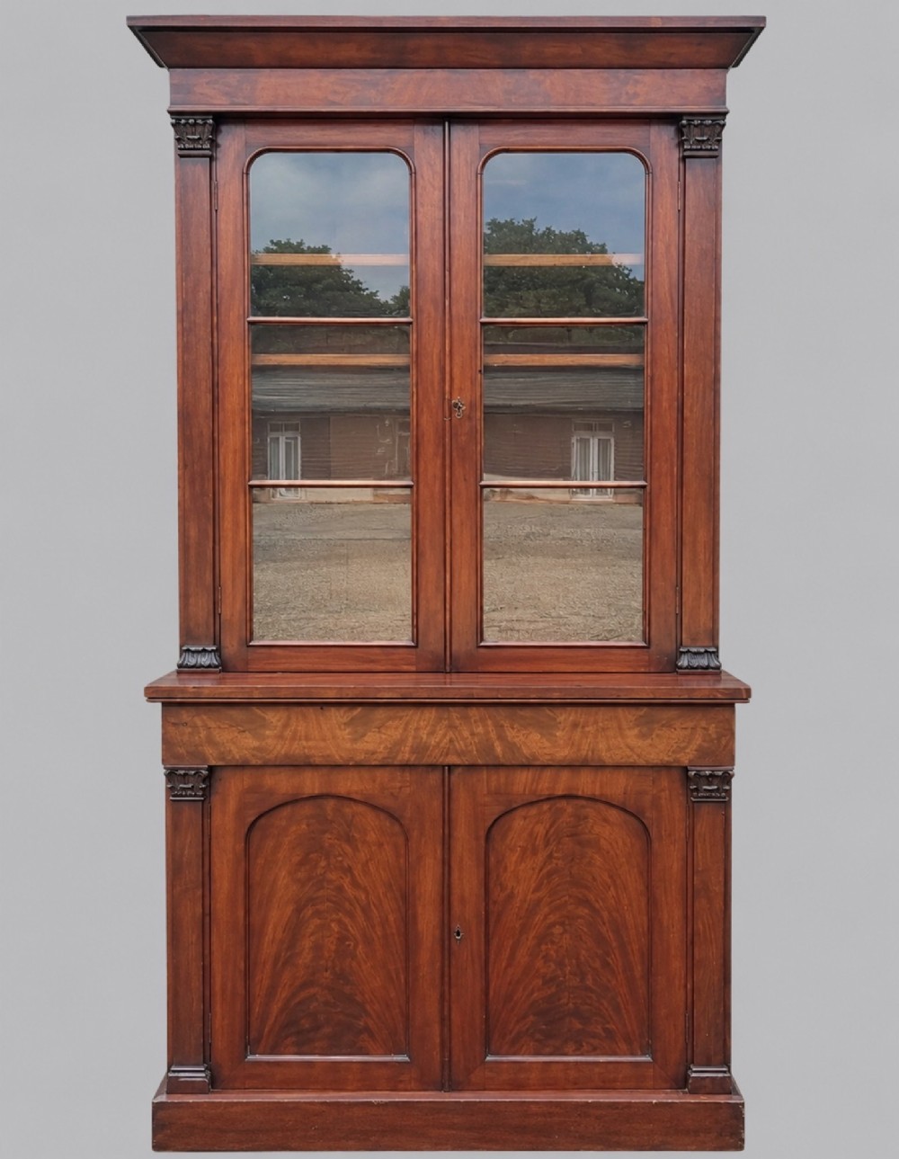 An early Victorian mahogany two-height library bookcase featuring upper glazed doors, flame mahogany lower panels, and carved pilasters.