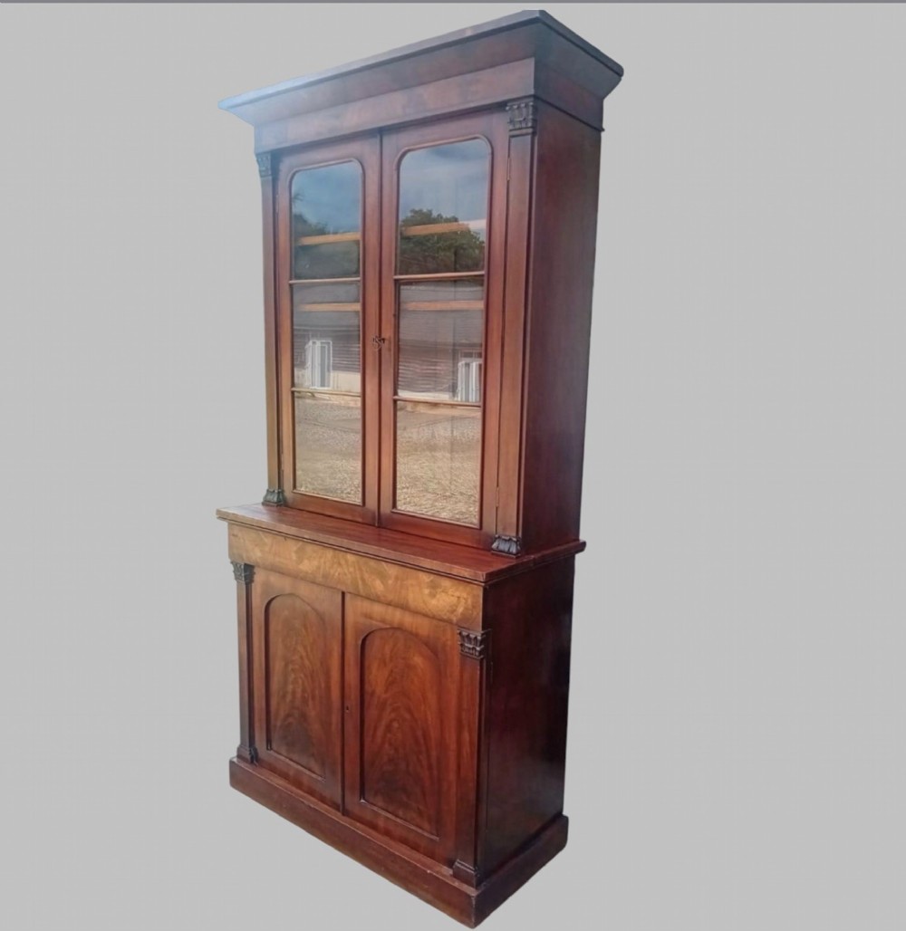 An early Victorian mahogany two-height library bookcase featuring upper glazed doors, flame mahogany lower panels, and carved pilasters. View II