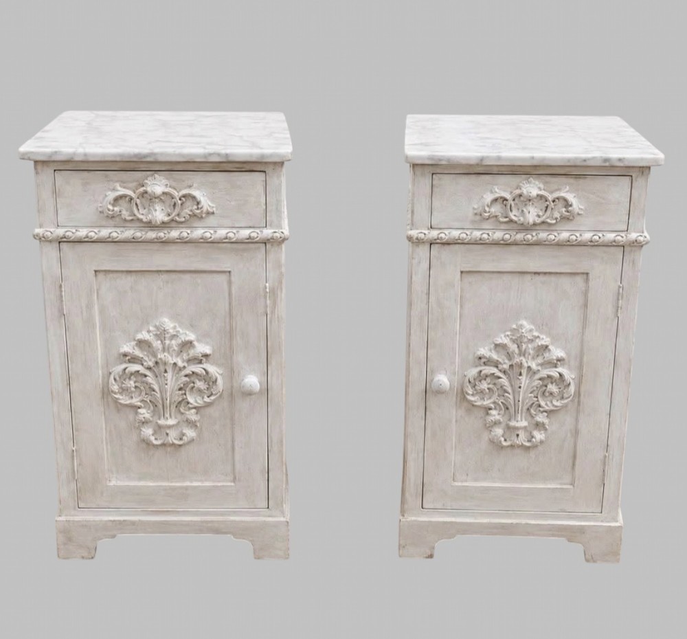 A pair of French off-white painted bedside cabinets featuring fitted marble tops, single drawers, and cupboard doors with central carved foliate relief motifs.