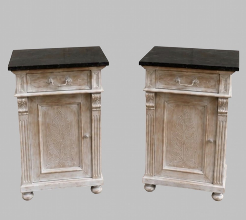 A pair of off-white painted wooden bedside cabinets with dark marble tops, single drawers, fluted pilasters, and carved leaf panels on the doors.