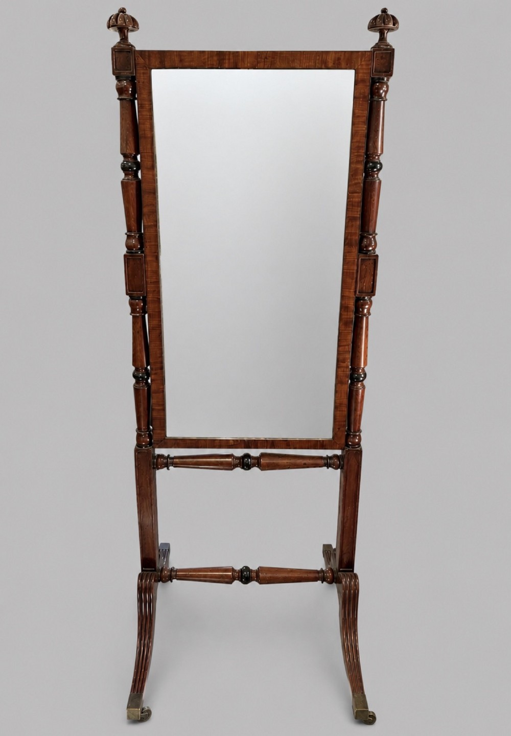 A nineteenth-century flame mahogany cheval mirror featuring turned side uprights, top finials, reeded splayed legs, and brass castors.