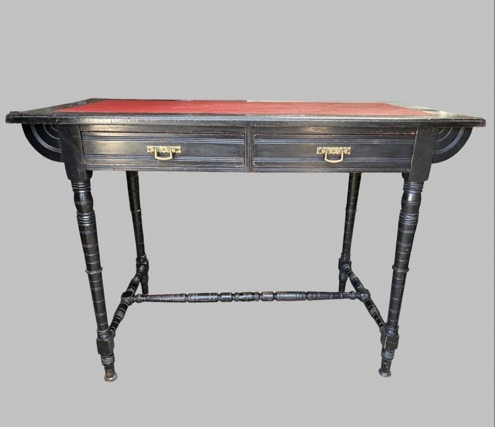 A late Victorian ebonised writing desk featuring a red leather top insert, two drawers with brass handles, turned legs, and an H-stretcher.