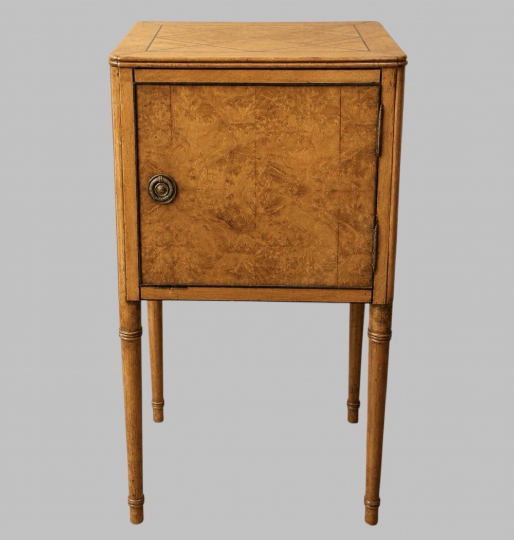 An Art Deco burr walnut bedside cabinet with a single door, circular brass handle, geometric inlaid top, and four slender tapered legs.