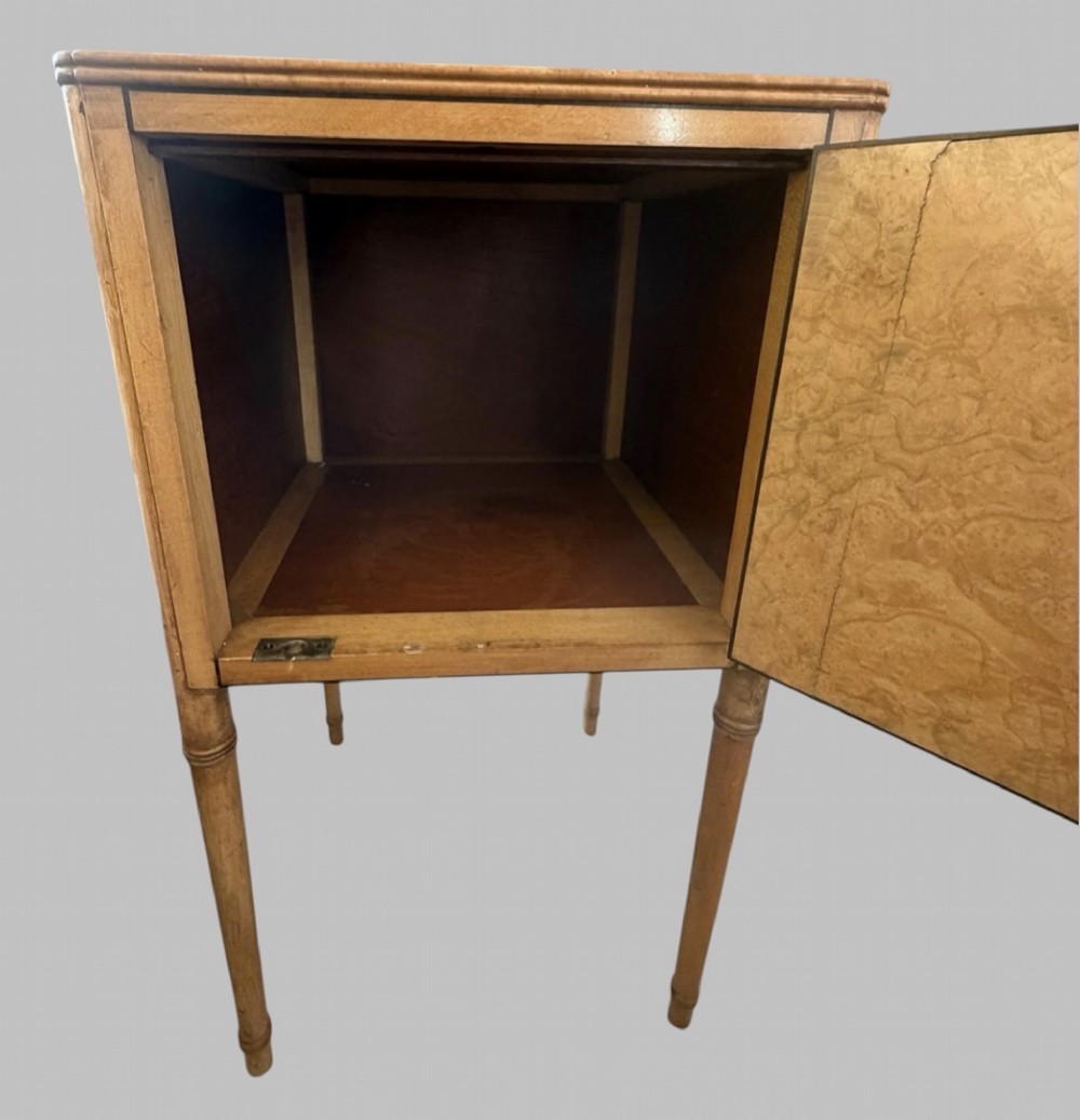 An Art Deco burr walnut bedside cabinet with a single door, circular brass handle, geometric inlaid top, and four slender tapered legs. View II