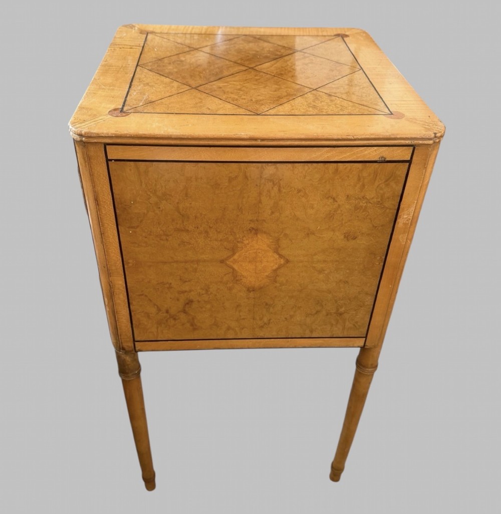 An Art Deco burr walnut bedside cabinet with a single door, circular brass handle, geometric inlaid top, and four slender tapered legs. View III