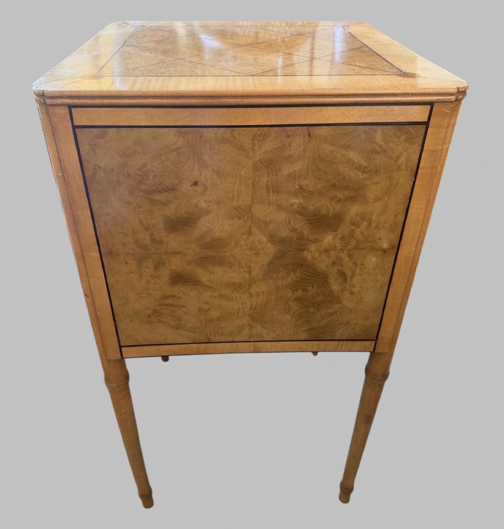 An Art Deco burr walnut bedside cabinet with a single door, circular brass handle, geometric inlaid top, and four slender tapered legs. View IV