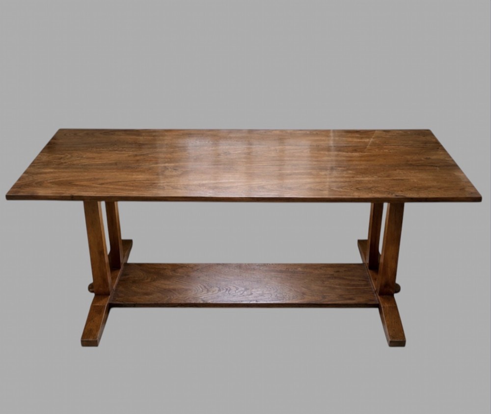 An Arts and Crafts solid oak dining table featuring a rectangular top, twin upright trestle end supports, and a low solid wooden stretcher shelf.