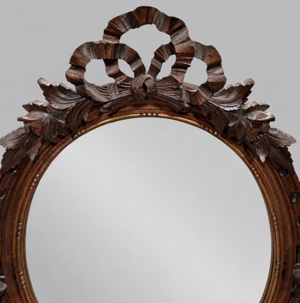 A late 19th-century oval wall mirror featuring a carved wooden frame topped with a sculpted ribbon cresting and floral motifs. View II