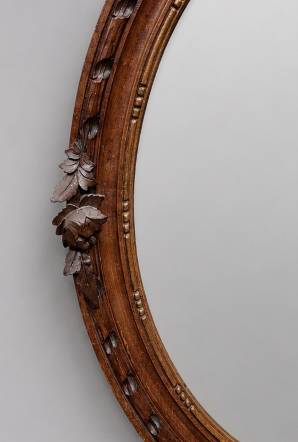 A late 19th-century oval wall mirror featuring a carved wooden frame topped with a sculpted ribbon cresting and floral motifs. View IV