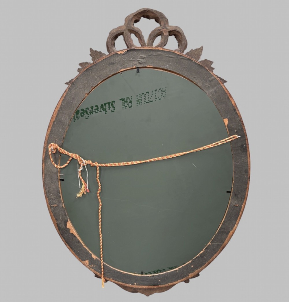 A late 19th-century oval wall mirror featuring a carved wooden frame topped with a sculpted ribbon cresting and floral motifs. View V