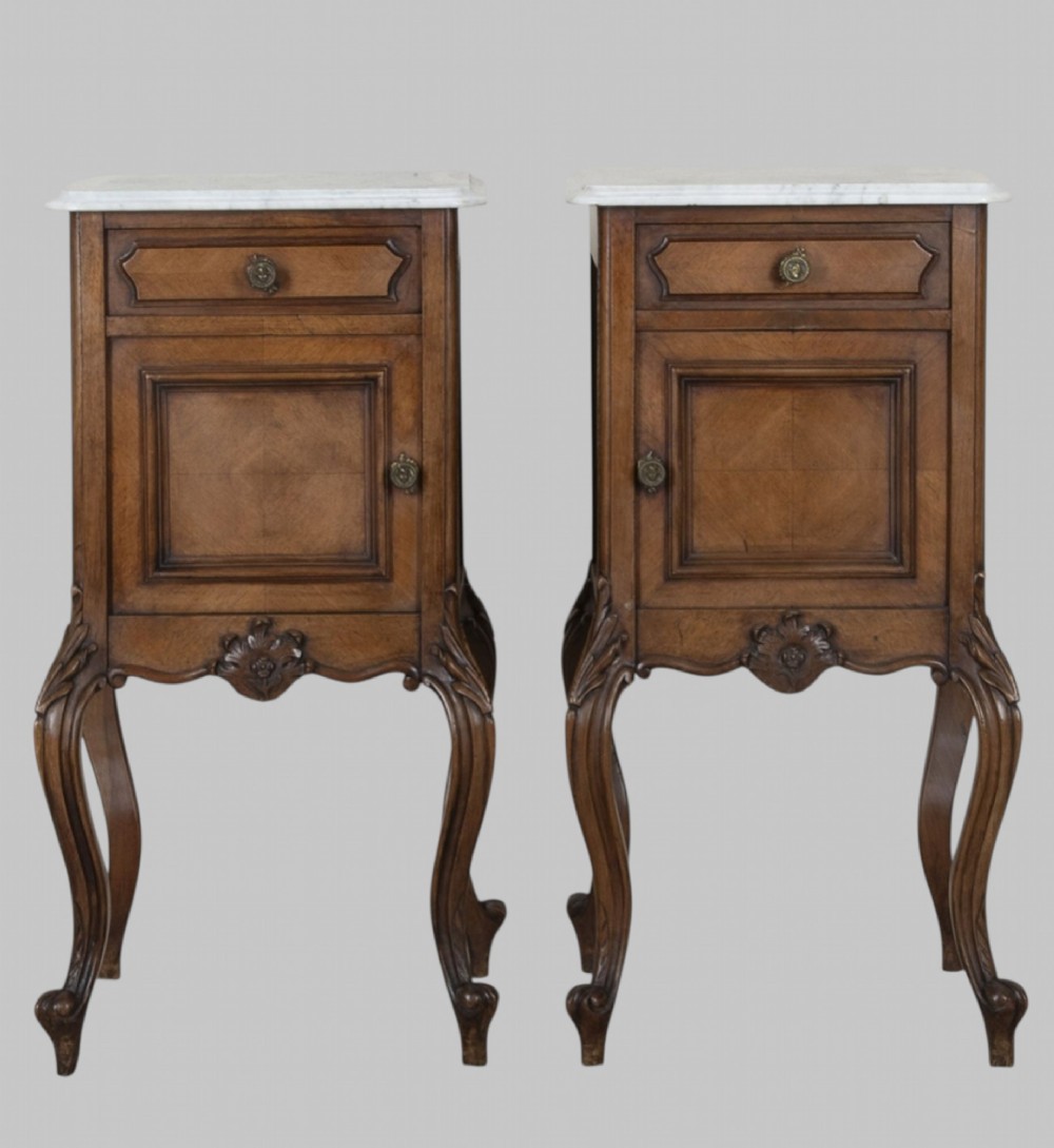A pair of French walnut bedside cabinets featuring white marble tops, single drawers over cupboard doors, carved aprons, and slender cabriole legs.