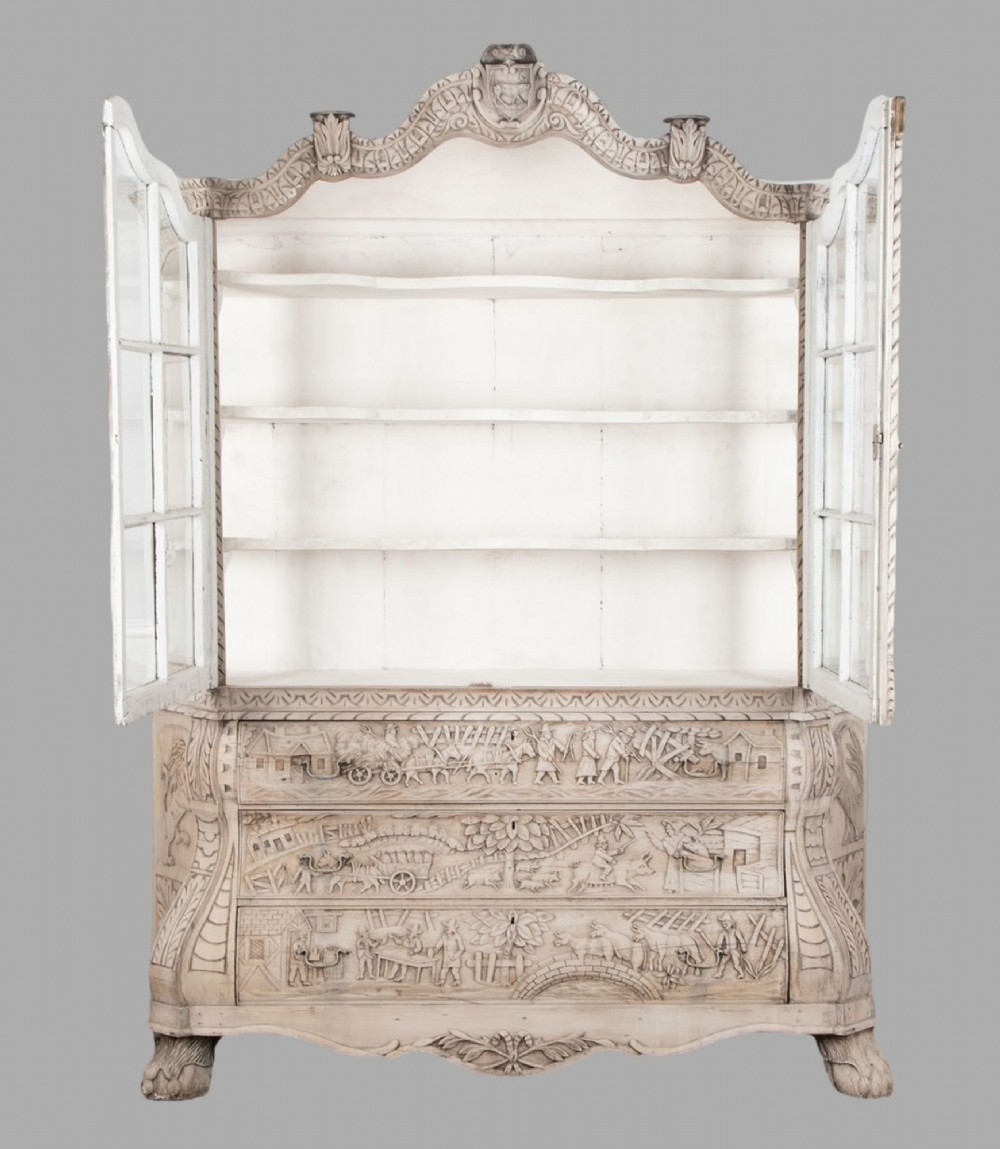 A tall nineteenth-century Dutch carved pale oak display cabinet featuring a shaped crest, multi-pane glazed upper doors, and a bombe chest base with three relief-carved drawers on paw feet. View II