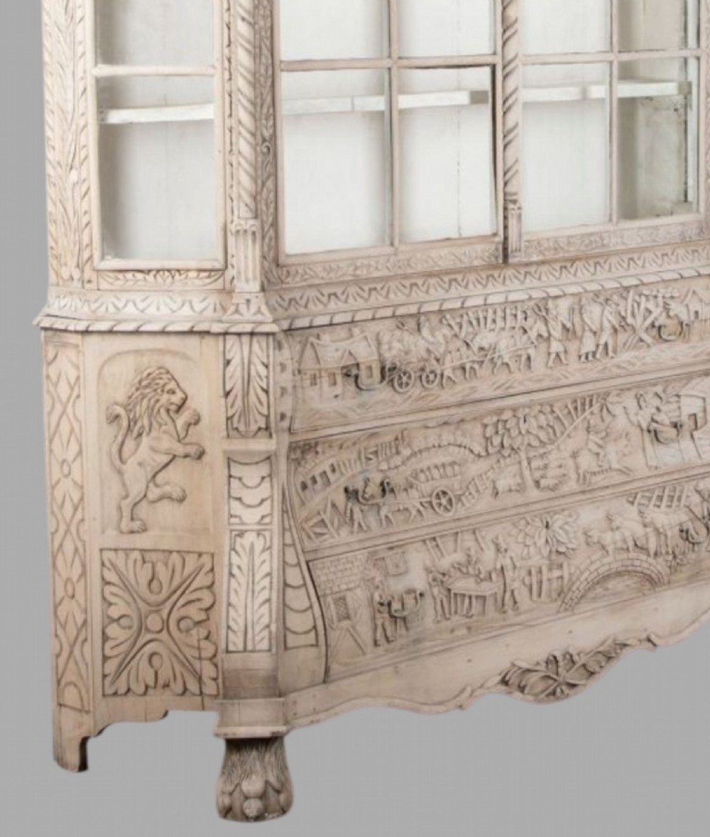A tall nineteenth-century Dutch carved pale oak display cabinet featuring a shaped crest, multi-pane glazed upper doors, and a bombe chest base with three relief-carved drawers on paw feet. View VII