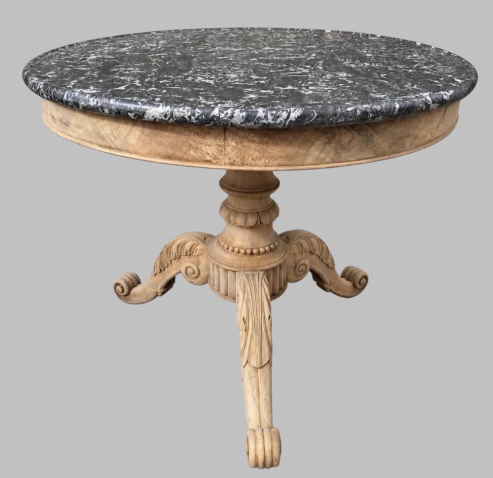 A circular antique French gueridon table featuring a dark grey mottled marble top supported by a carved bleached walnut pedestal and tripod scrolled legs.