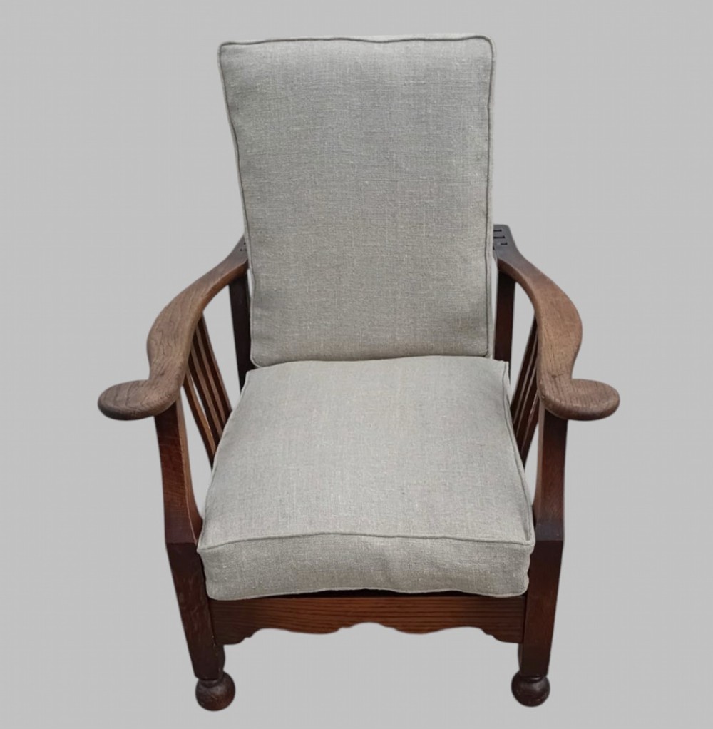 An Arts and Crafts dark oak reclining armchair with wide curved armrests and light grey fabric back and seat cushions.