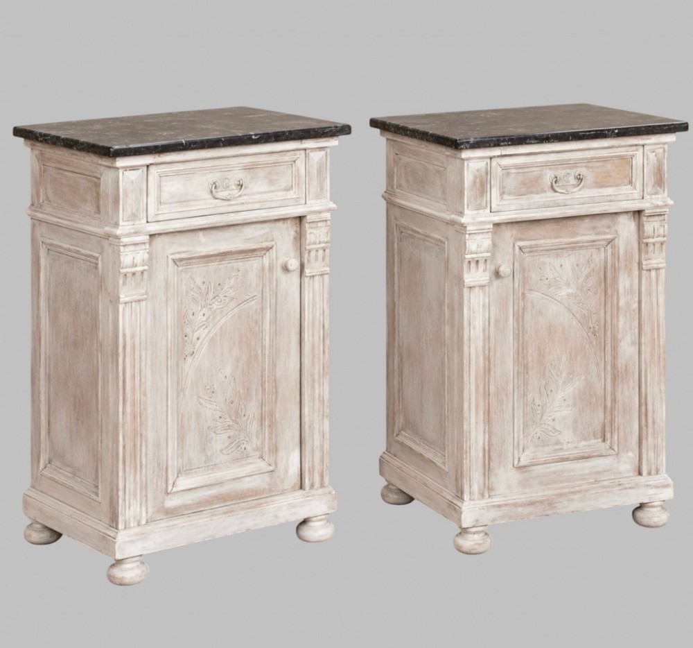 A matching pair of mid-nineteenth-century off-white painted wooden bedside cabinets featuring dark marble tops, single drawers, fluted side pilasters, carved floral door panels, and turned bun feet.
