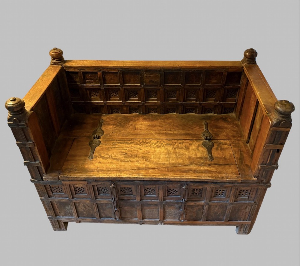 A carved dark hardwood storage bench with a hinged lid seat, geometric panelled backrest, corner post finials, and iron fittings. View II