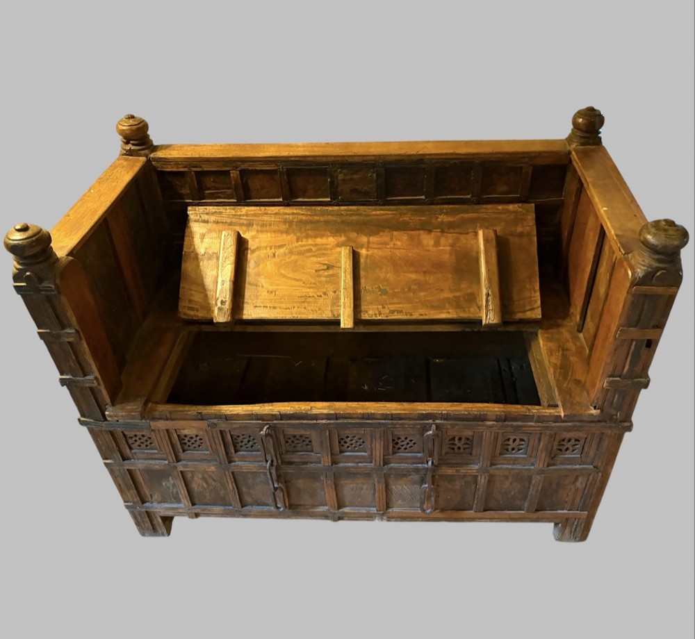 A carved dark hardwood storage bench with a hinged lid seat, geometric panelled backrest, corner post finials, and iron fittings. View III