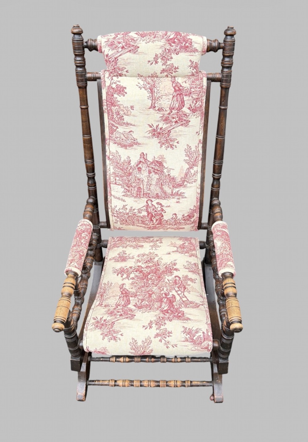 An antique turned wood rocking chair featuring dark timber spindle armrests and frame with cream fabric upholstery displaying red figurative pattern work. View II